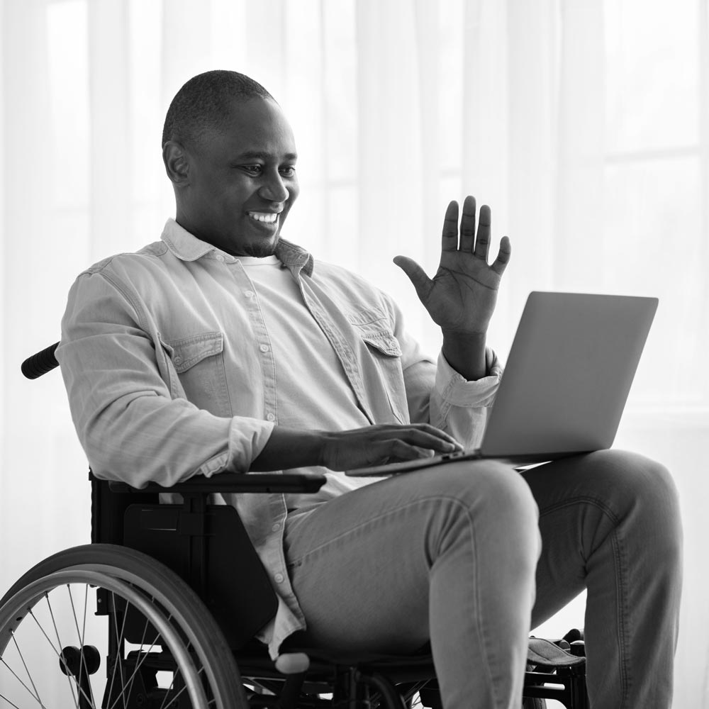 A man in a wheelchair waving hello to someone on a virtual call on his laptop.